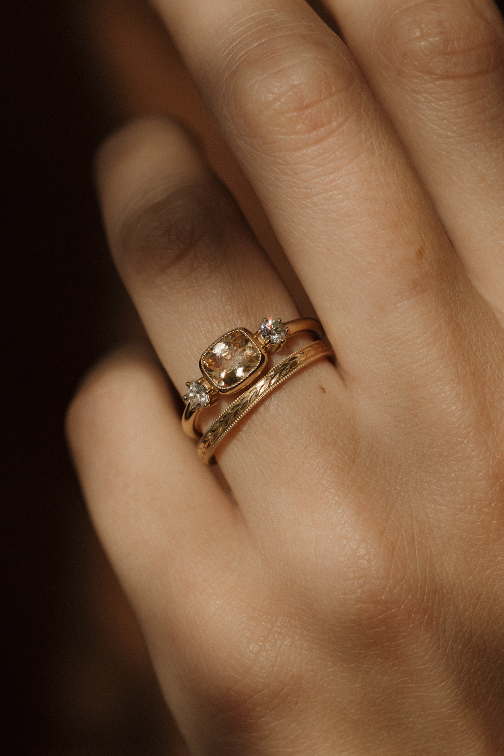 Close view of a hand wearing a yellow gold engagement ring with an east-west cushion cut peach sapphire and two old cut diamonds, stacked with a slim hand-engraved wedding band edged in milgrain.