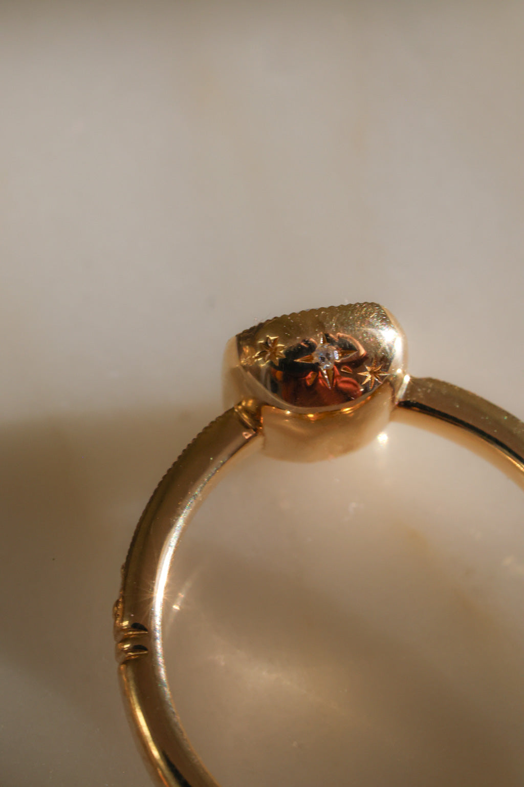 Close view of the side and underside of a yellow gold engagement ring's bezel cup, showing a star-set diamond between two engraved stars, the milgrain edge of the band, and a grooved band detail, on a reflective cream marble surface in warm light.