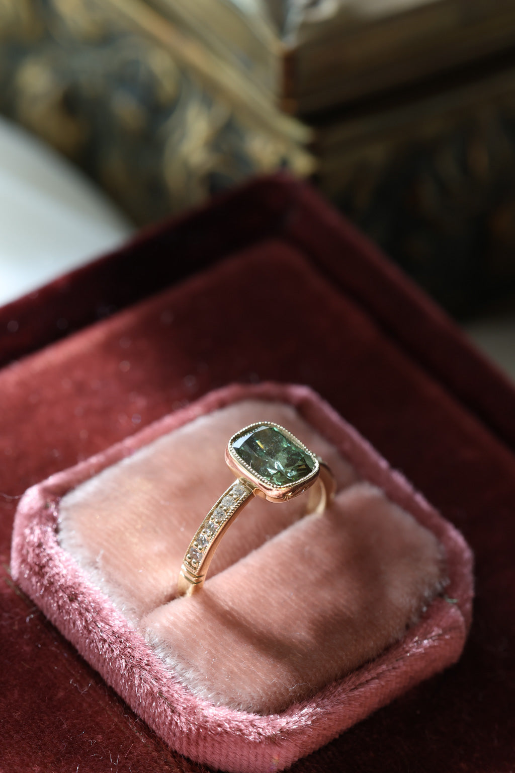 Angled view of a yellow gold engagement ring with a cushion cut green diamond in a slim milgrain bezel and a diamond-set band, standing in a blush velvet ring box on burgundy velvet with an antique-style brass jewelry box blurred behind.