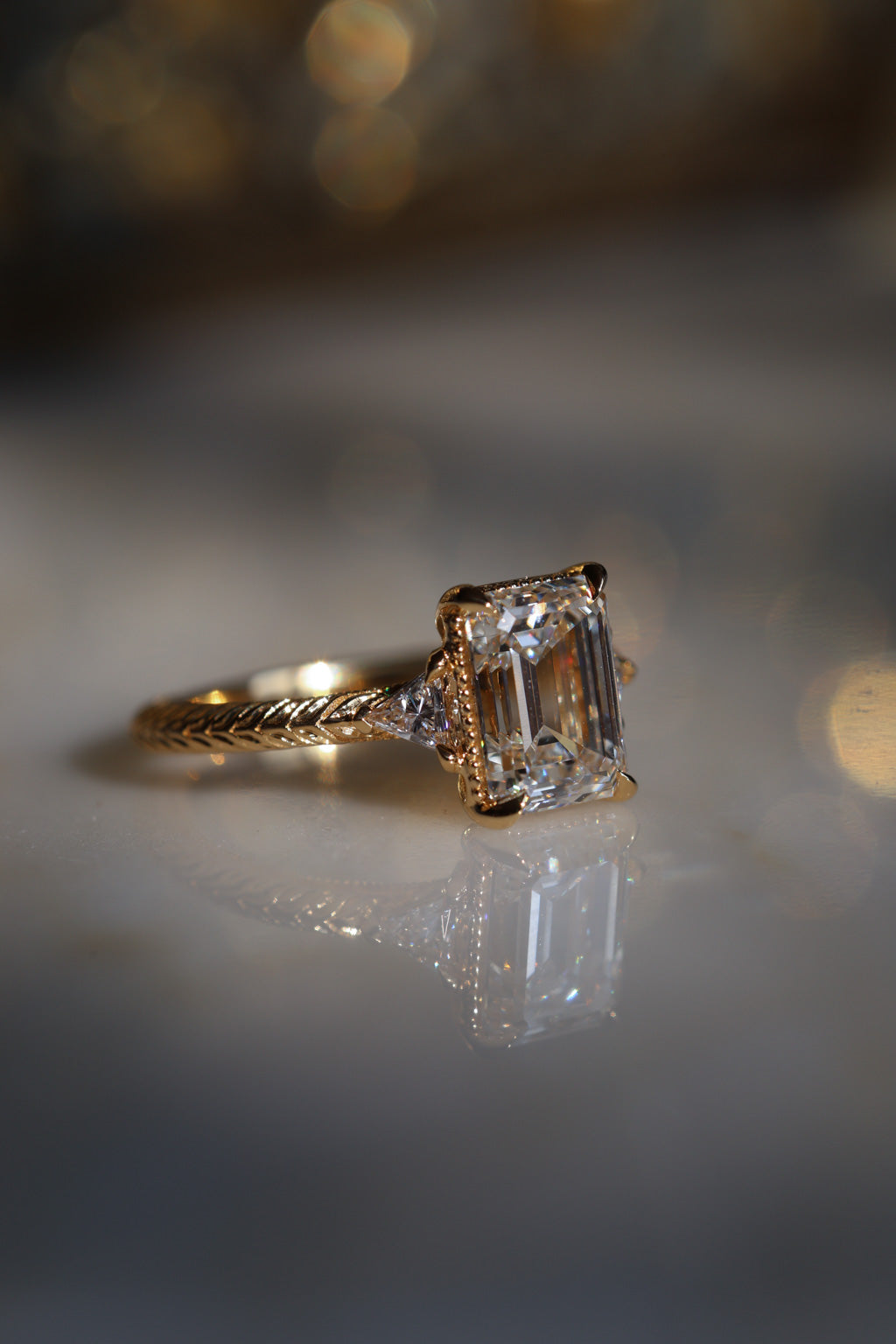 A picture of an engagement ring resting on a marble surface with a soft blur around it. The ring is a yellow gold hand-engraved band and milgrain details. It has an emerald cut diamond in a three stone setting and the accent diamonds are triangle cut. 