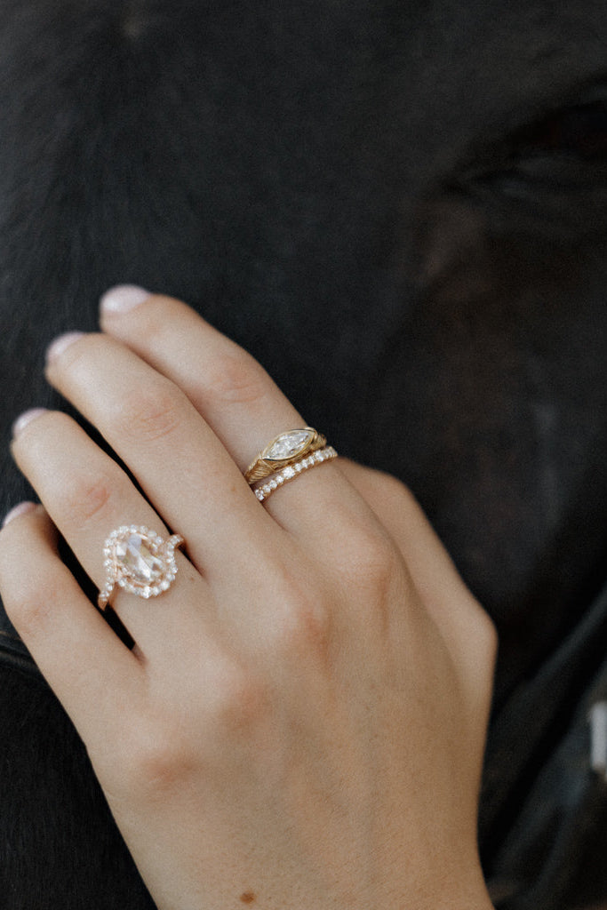 Photo of a hand resting against a dark brown horse's coat, wearing an engagement ring and a thin diamond eternity band on the middle finger, and another ring on the ring finger. The engagement ring is a chunky yellow gold hand-engraved band with a marquise cut diamond set east-west in a bezel. The other ring is rose gold with a rose cut diamond in a halo.