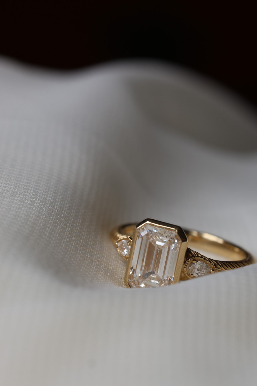 An engagement ring is laying on white fabric, the bottom corner is partially obstructed by the fabric. The ring is yellow gold with a hand engraved detail. The center stone is a bezel set emerald cut diamond, with two pear shaped diamond accents that have a milgrain border. 