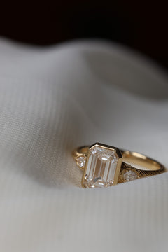 An engagement ring is laying on white fabric, the bottom corner is partially obstructed by the fabric. The ring is yellow gold with a hand engraved detail. The center stone is a bezel set emerald cut diamond, with two pear shaped diamond accents that have a milgrain border. 