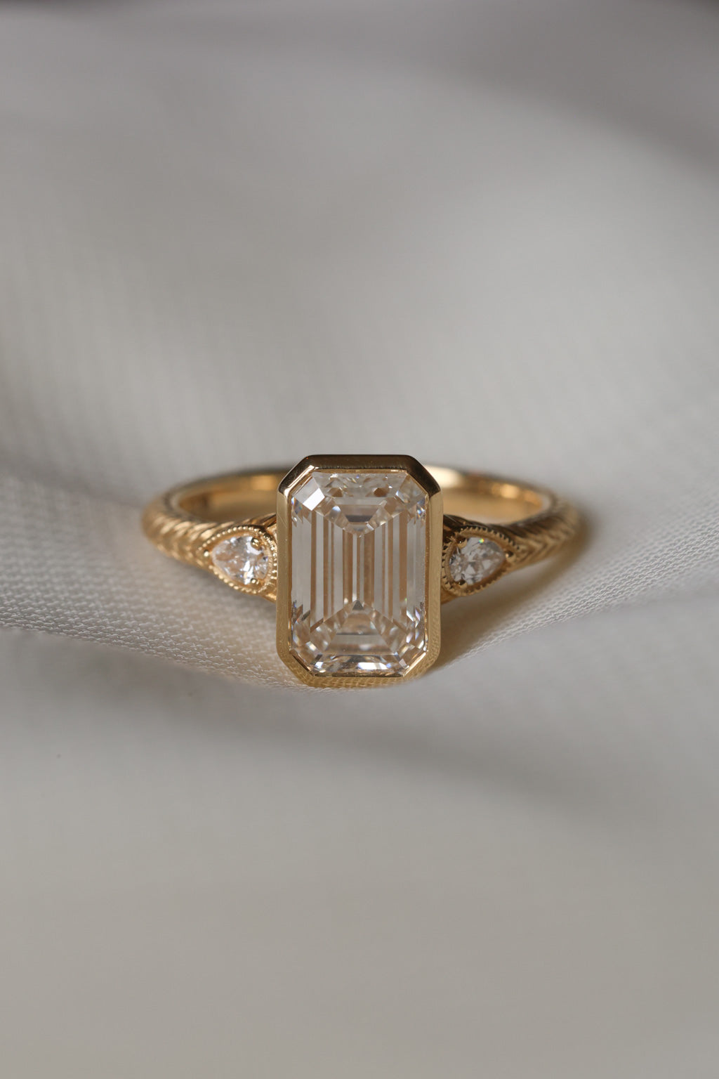A yellow gold hand engraved engagement ring sits on white fabric. The ring has an emerald cut diamond in a bezel setting with two pear cut side stones with a milgrain border. 