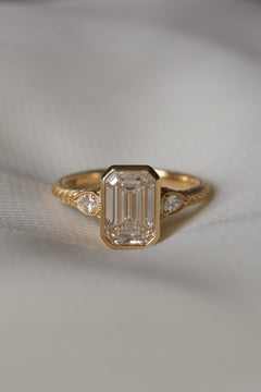 A yellow gold hand engraved engagement ring sits on white fabric. The ring has an emerald cut diamond in a bezel setting with two pear cut side stones with a milgrain border. 