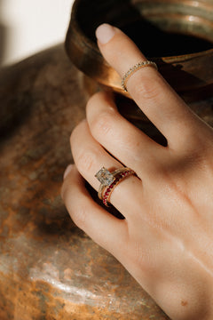 On-hand view of a yellow gold engagement ring with an antique emerald cut diamond on a hand-engraved band, stacked with the Moonryvr French Pave Ruby Band, with a diamond band on the index finger, the hand resting on a weathered copper vessel.