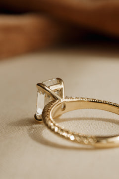 Back view of a yellow gold engagement ring showing the claw prongs and gallery beneath an antique emerald cut diamond and the hand-engraved band with milgrain edges, on cream woven linen against a warm brown background.