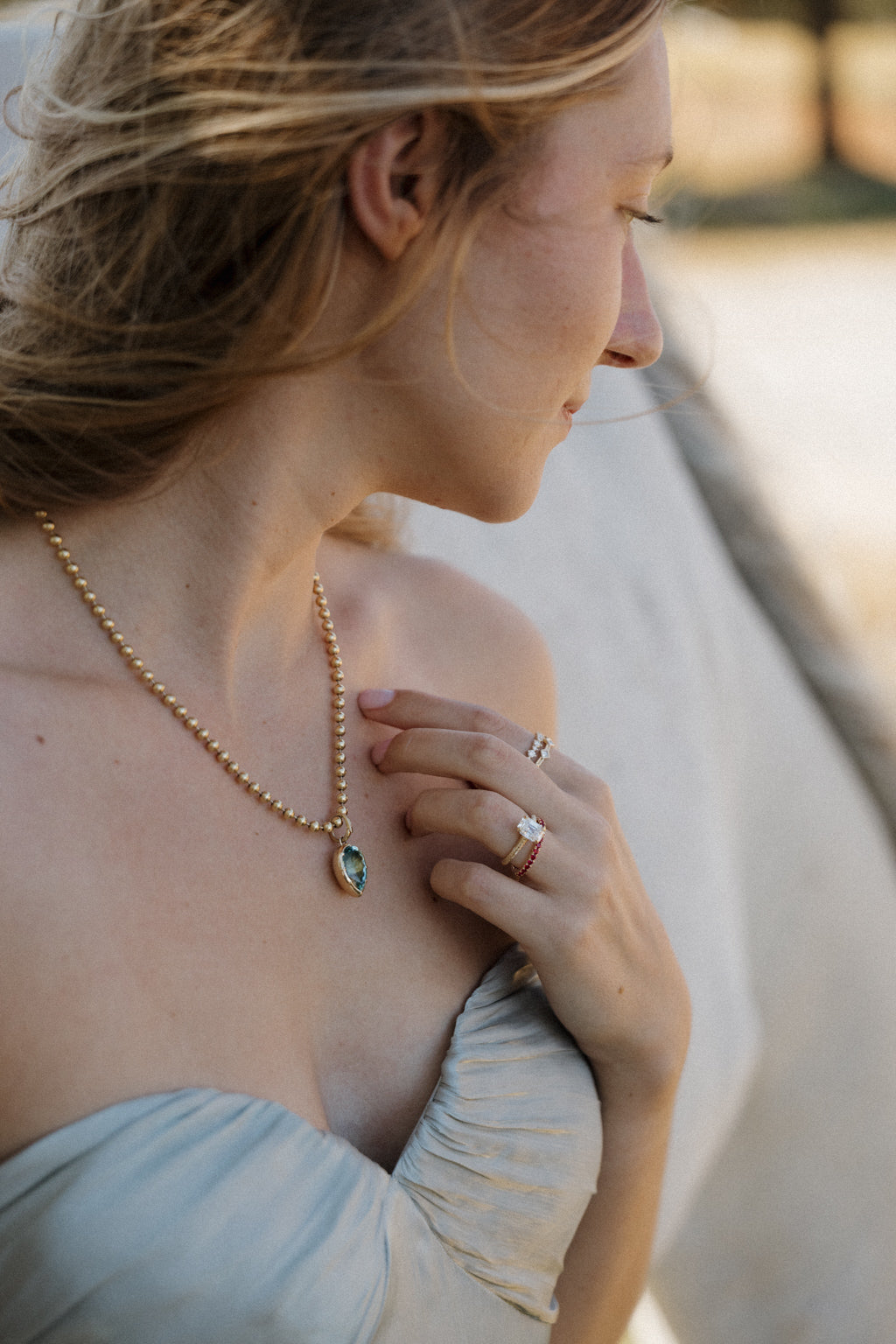 On-hand view of a yellow gold engagement ring with an antique emerald cut diamond stacked with the Moonryvr French Pave Ruby Band, with the Square Diamond Band and Alternating Size Round Diamond Band on the index finger. The hand rests at the shoulder of a pale blue off-shoulder dress beside a gold bead chain necklace with a bezel-set pear-shaped blue-green pendant, a white horse blurred behind.