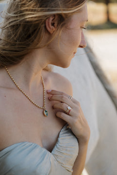 On-hand view of a yellow gold engagement ring with an antique emerald cut diamond stacked with the Moonryvr French Pave Ruby Band, with the Square Diamond Band and Alternating Size Round Diamond Band on the index finger. The hand rests at the shoulder of a pale blue off-shoulder dress beside a gold bead chain necklace with a bezel-set pear-shaped blue-green pendant, a white horse blurred behind.