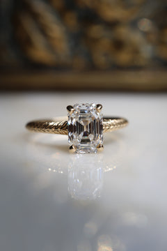 Close front view of a yellow gold solitaire engagement ring with an antique emerald cut diamond held by four claw prongs on a hand-engraved band, on a reflective cream marble surface with an antique-style brass jewelry box blurred behind.