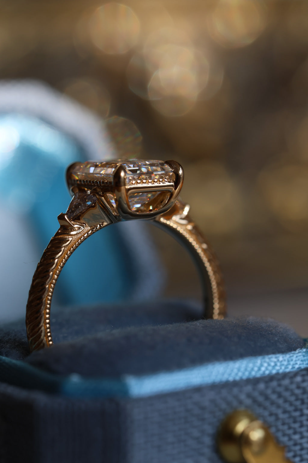 Close up image of an open blue ring box with a side view of an engagement ring. The ring is a yellow gold hand-engraved band and milgrain details. It has an emerald cut diamond in a three stone setting and the accent diamonds are triangle cut. It has an open gallery with four prongs, and there is a milgrain detail on the edge of the ring. 