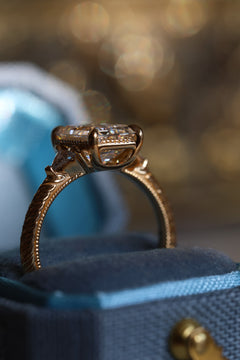 Close up image of an open blue ring box with a side view of an engagement ring. The ring is a yellow gold hand-engraved band and milgrain details. It has an emerald cut diamond in a three stone setting and the accent diamonds are triangle cut. It has an open gallery with four prongs, and there is a milgrain detail on the edge of the ring. 