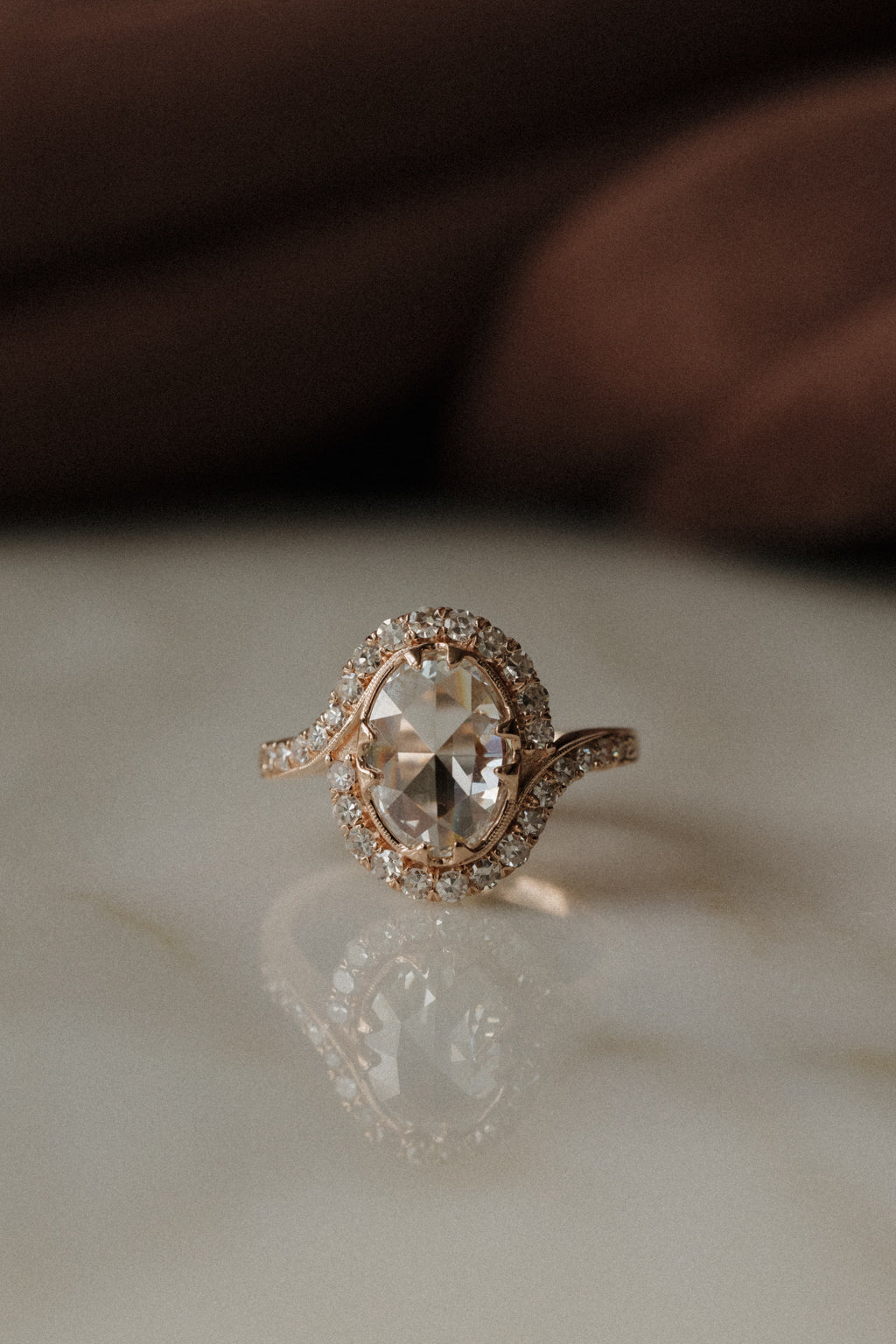 A front view of a rose gold engagement ring sitting on a marble surface. The ring has an oval shape rose cut diamond center stone with a single cut diamond bypass halo. The oval has four double prongs and the band has a small engraving where the halo ends.