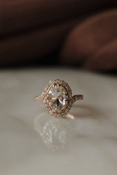 A front view of a rose gold engagement ring sitting on a marble surface. The ring has an oval shape rose cut diamond center stone with a single cut diamond bypass halo. The oval has four double prongs and the band has a small engraving where the halo ends.