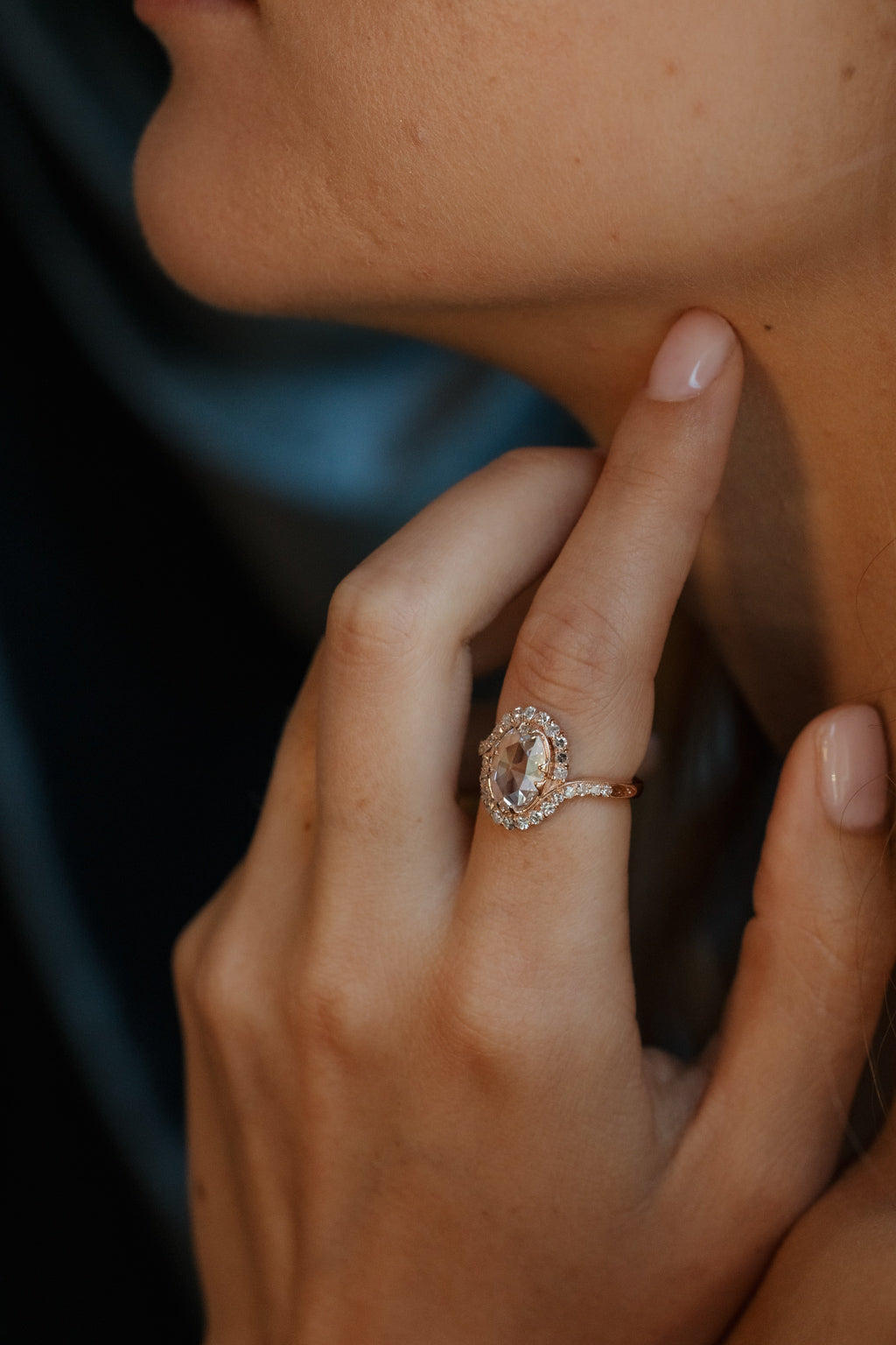 Close up of a side profile view of a woman, her hand is touching her neck and wearing a rose gold engagement ring. The engagement ring is an oval shape rose cut diamond with a single cut diamond bypass halo, the band has engravings where the halo stops and there are four double prongs in a compass setting around the center stone.