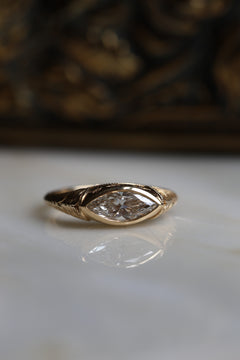 Close up photo of an engagement ring standing on a white reflective surface, with a gilded frame out of focus behind it. The ring is a chunky yellow gold hand-engraved band. It has a marquise cut diamond set east-west in a bezel with milgrain along the edge, and the ring is reflected in the surface below.
