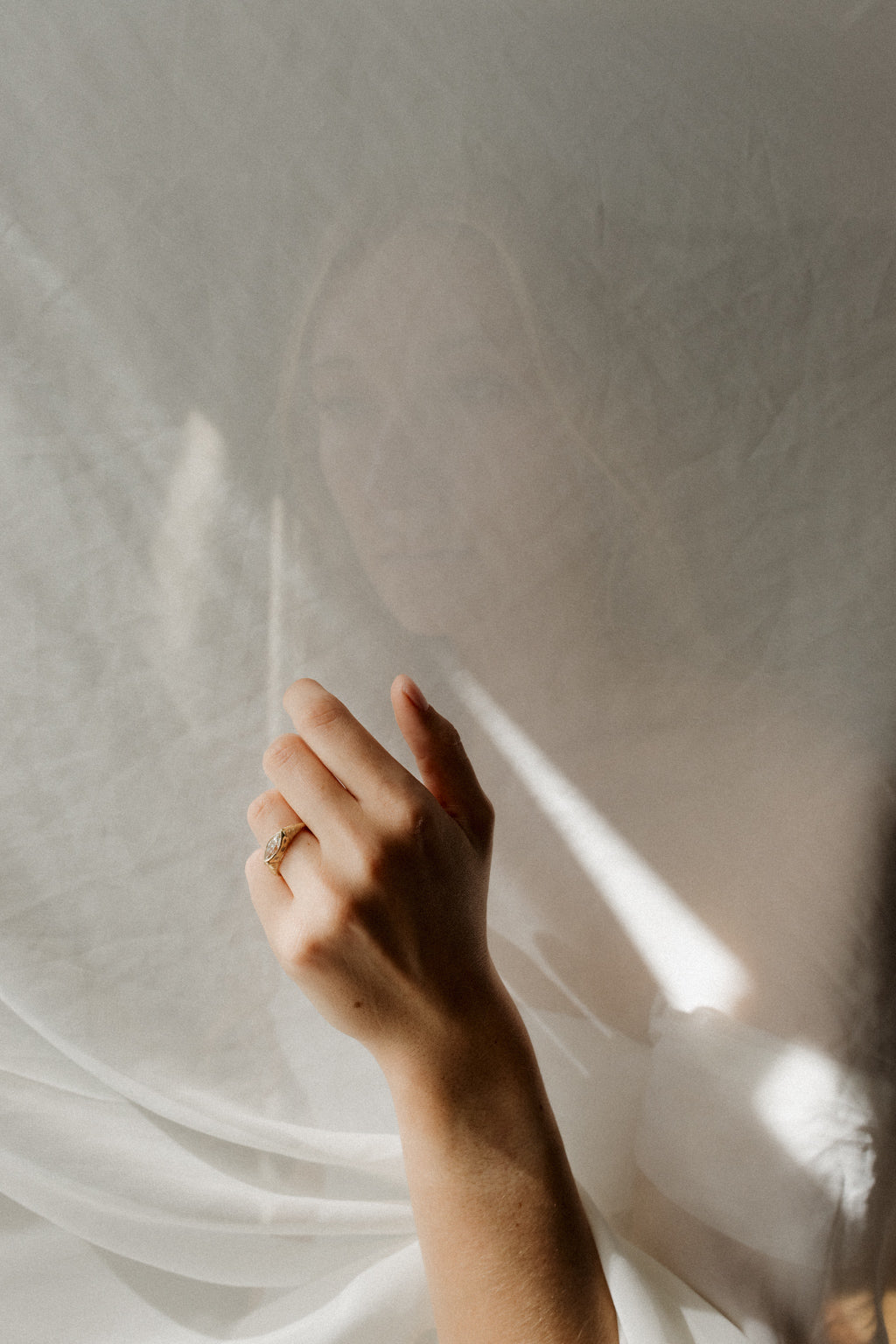 Photo of a hand and forearm reaching up against a sunlit white curtain, wearing an engagement ring on the ring finger, with a shadow falling across the fabric behind. The ring is a chunky yellow gold hand-engraved band with a marquise cut diamond set east-west in a bezel.