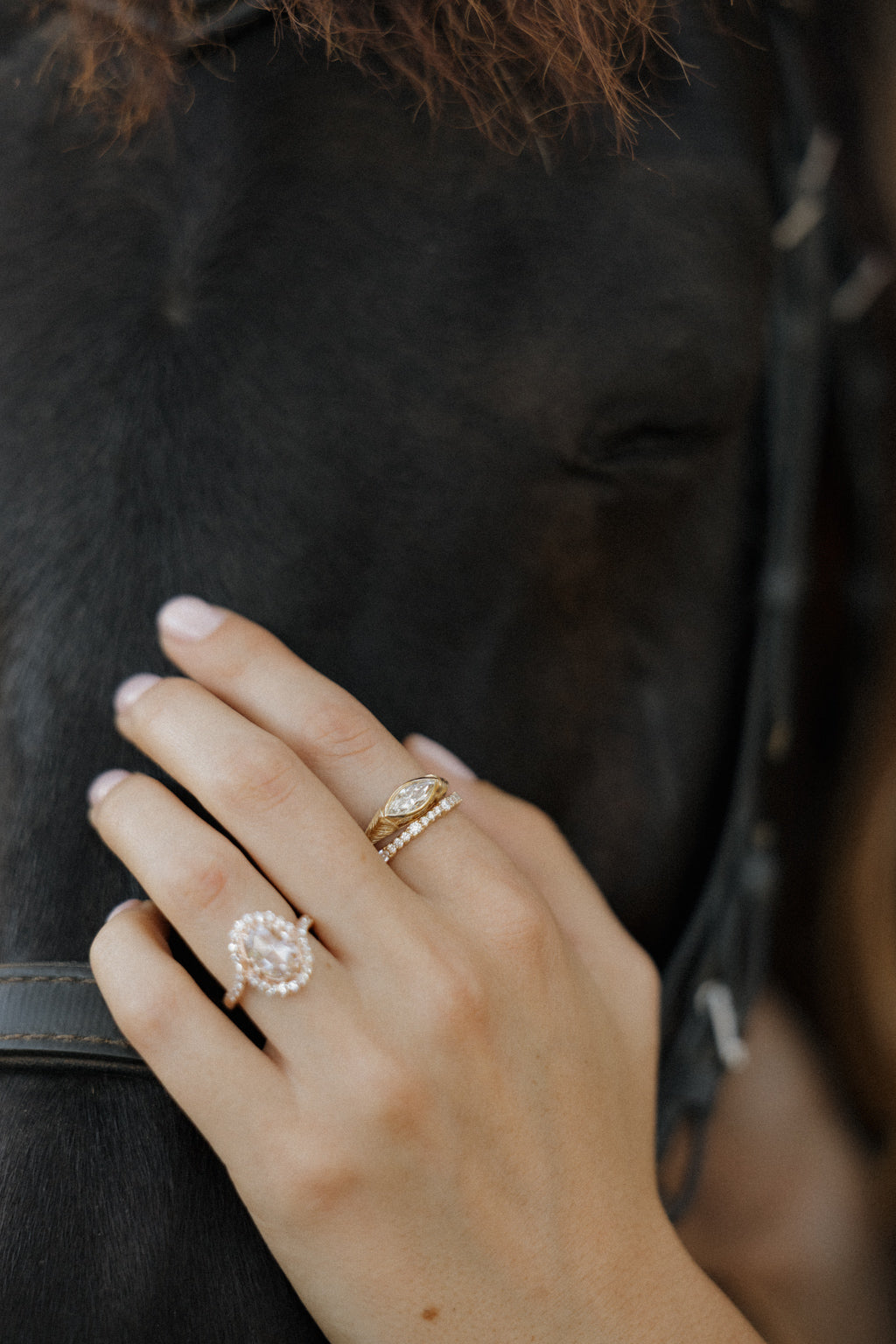Photo of a hand resting against a dark brown horse's face beside a leather bridle, with the horse's mane out of focus above. The hand wears an engagement ring and a thin diamond eternity band on the middle finger, and a rose gold ring with a rose cut diamond in a halo on the ring finger. The engagement ring is a chunky yellow gold hand-engraved band with a marquise cut diamond set east-west in a bezel.
