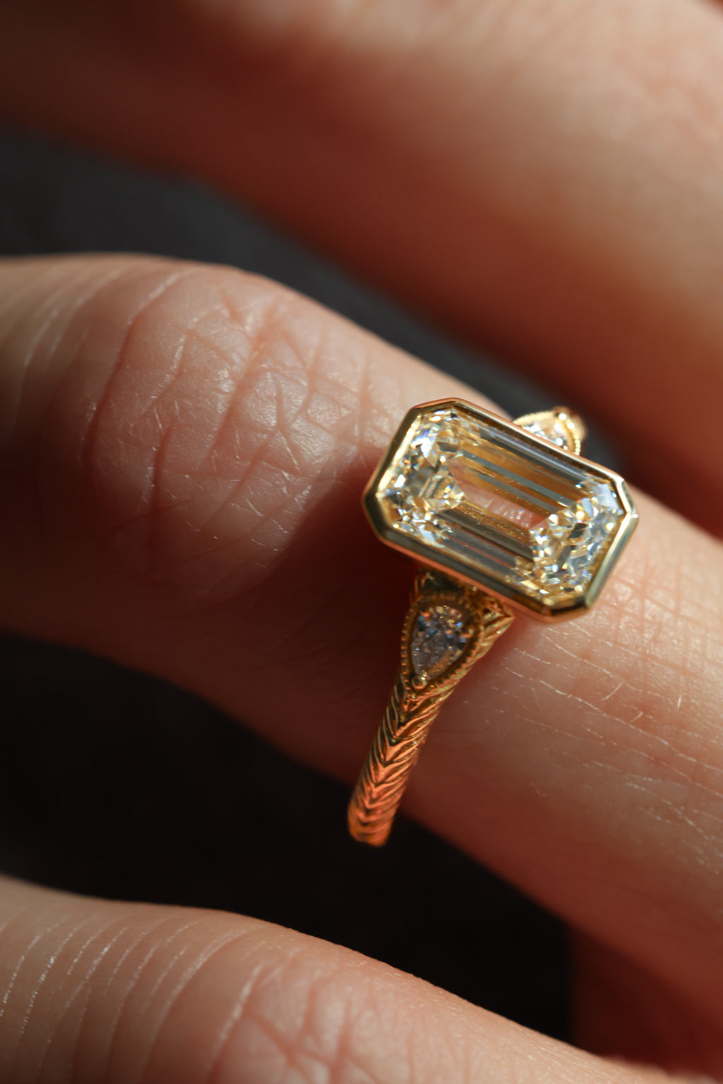 A close up of a finger wearing a three stone diamond engagement ring. The ring is yellow gold with a hand engraved band. The center stone is a bezel set emerald cut diamond with two pear shaped accent stones and a milgrain border.  