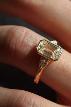 A close up of a finger wearing a three stone diamond engagement ring. The ring is yellow gold with a hand engraved band. The center stone is a bezel set emerald cut diamond with two pear shaped accent stones and a milgrain border.  