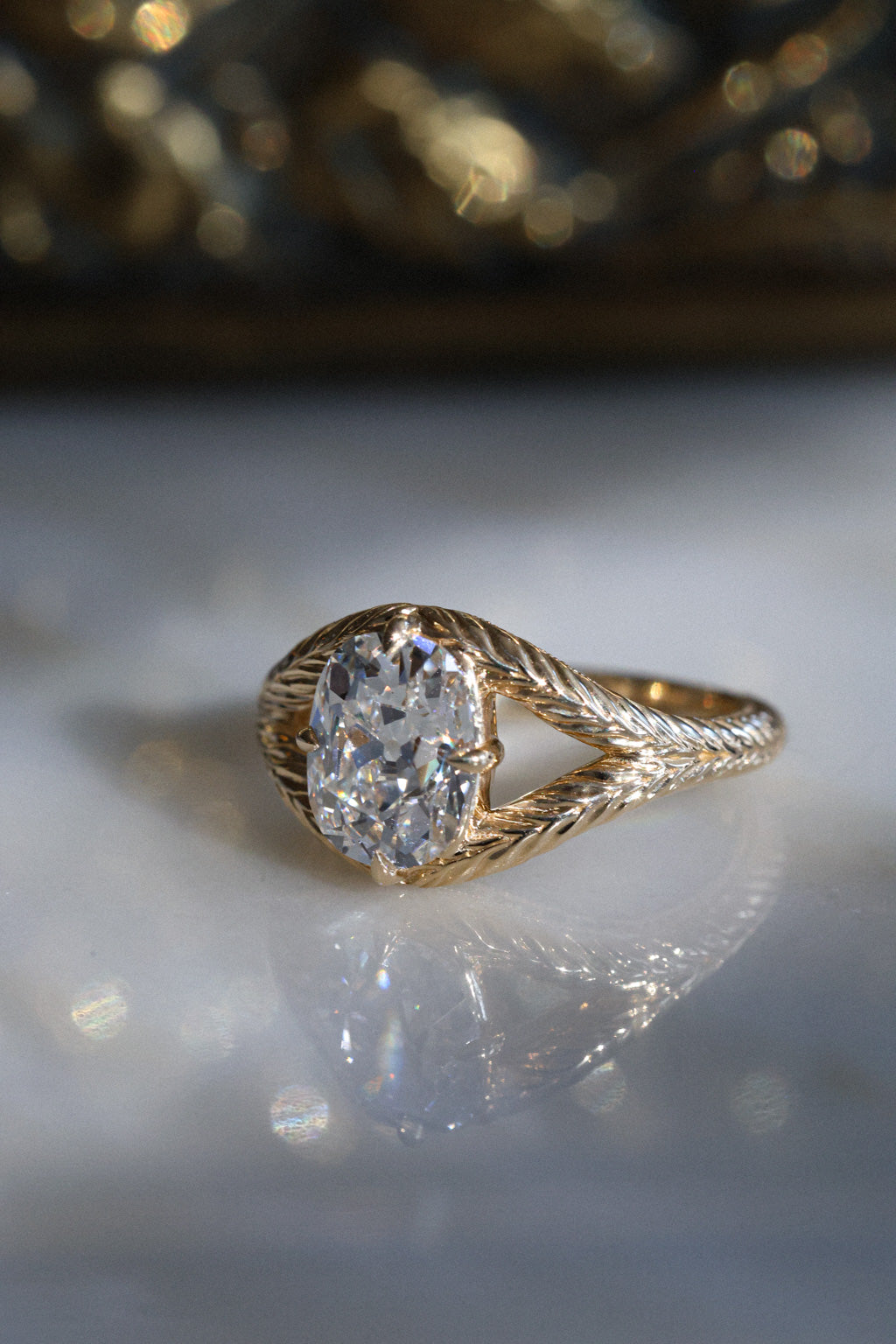 An engagement ring resting on a marble surface, the ring has a yellow gold old-world wheat hand-engraved band, a split shank, and four claw prongs in a compass setting around an old mine cut diamond.