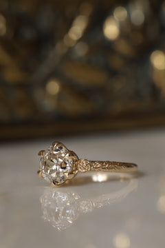 Angled side view of a yellow gold engagement ring with an old European cut diamond held by six claw prongs, a sculpted rose at the shoulder, and a vine motif along the band, on a reflective cream marble surface with an antique-style brass jewelry box blurred behind.