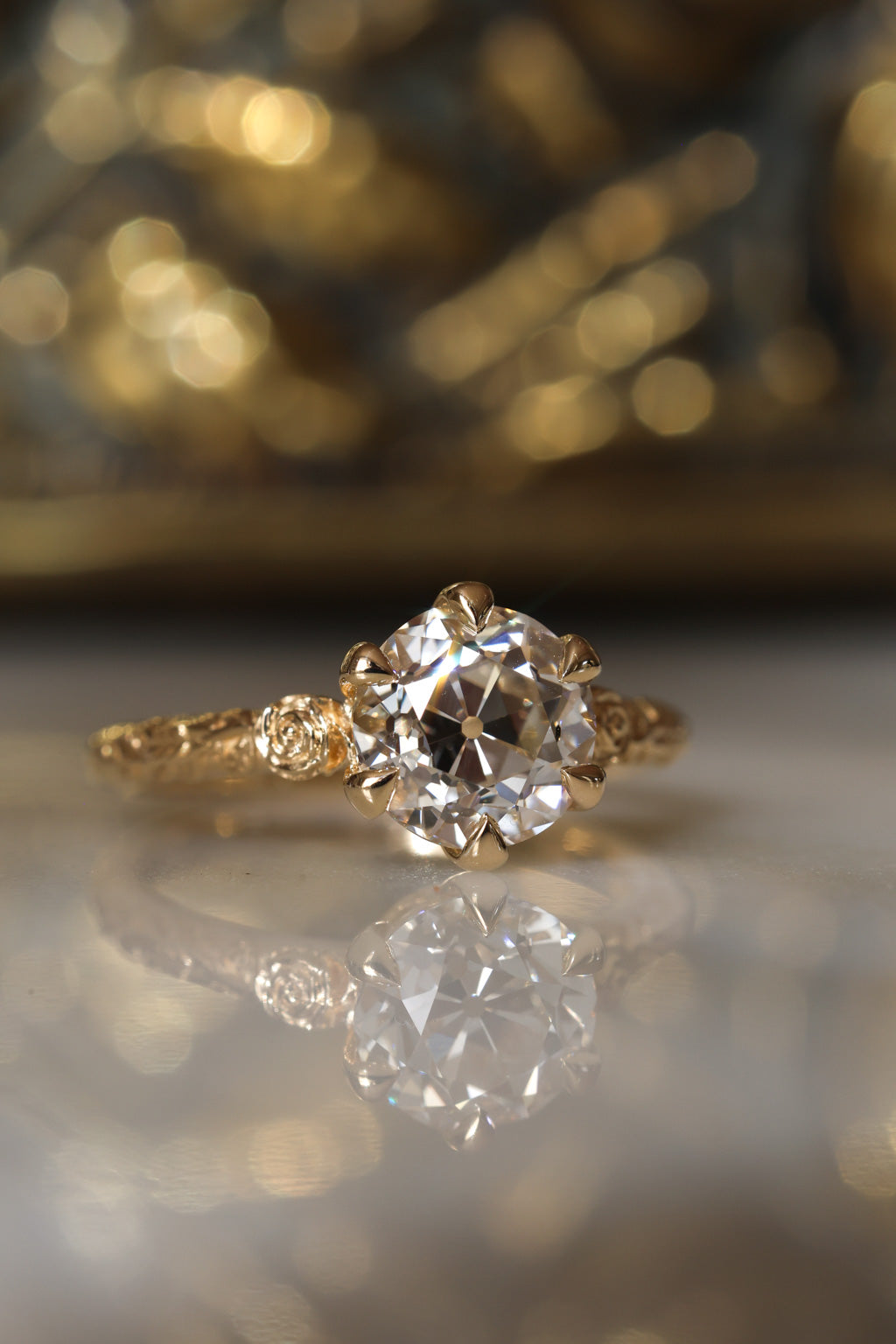 Front view of a yellow gold engagement ring with an old European cut diamond held by six claw prongs, flanked by sculpted roses on each shoulder and a vine motif along the band, on a reflective cream marble surface with an antique-style brass jewelry box blurred behind.