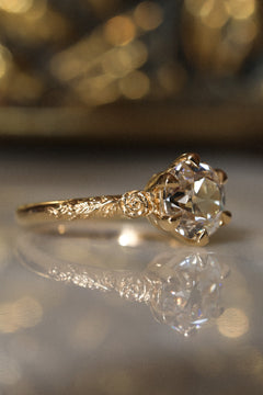 Angled side view of a yellow gold engagement ring with an old European cut diamond facing right in six claw prongs, a sculpted rose at the shoulder, and a vine motif along the band, on a reflective cream marble surface with an antique-style brass jewelry box blurred behind.