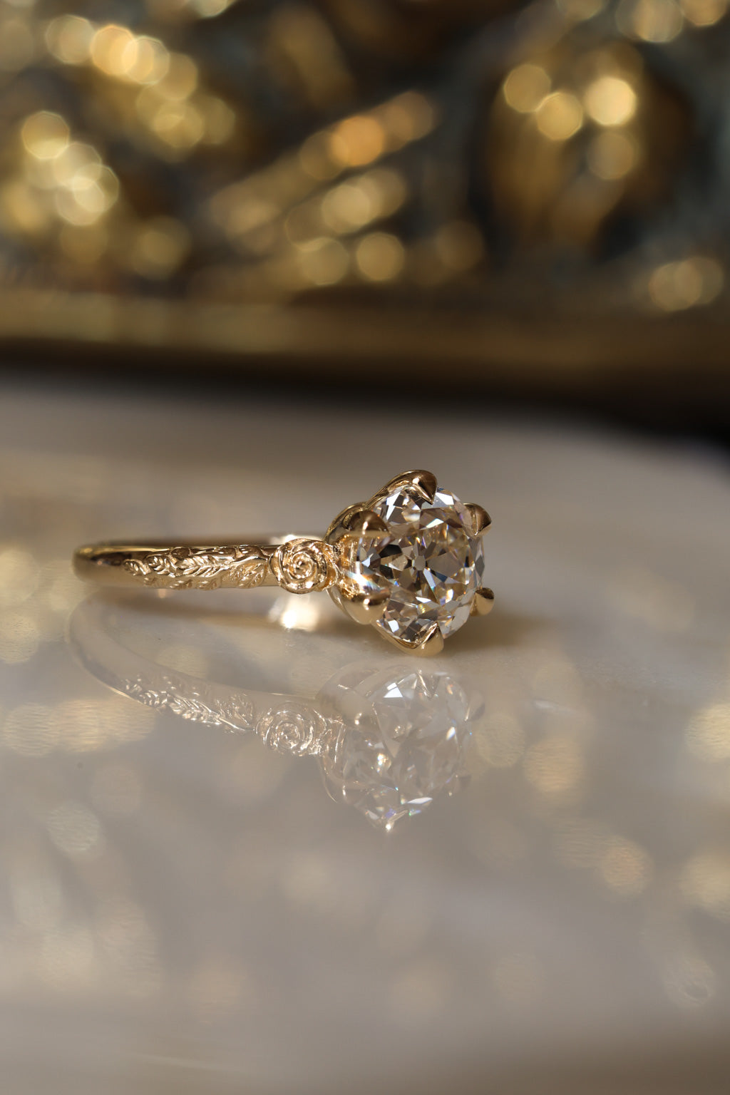 Angled side view of a yellow gold engagement ring with an old European cut diamond in six claw prongs and a sculpted rose and vine band, on a reflective cream marble surface with an antique-style brass jewelry box blurred behind.