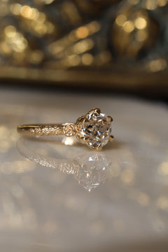 Angled side view of a yellow gold engagement ring with an old European cut diamond in six claw prongs and a sculpted rose and vine band, on a reflective cream marble surface with an antique-style brass jewelry box blurred behind.