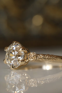 Close-up side view of a yellow gold engagement ring showing the six claw prongs around an old European cut diamond, a sculpted rose at the shoulder, and the vine motif along the band, on a reflective cream marble surface.