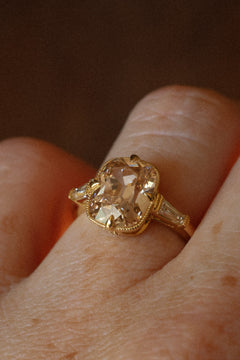 Close view of a hand wearing a yellow gold engagement ring with a champagne old mine cut diamond held in claw prongs and edged in milgrain, between two tapered baguette diamonds, against a dark background.