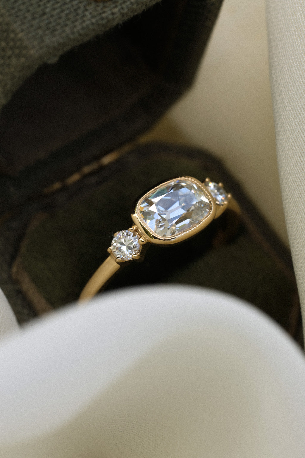 Close up photo of an engagement ring resting on the edge of an open antique jewelry box lined with dark velvet, with cream fabric in the foreground and gray-green cloth behind. The ring is a thin yellow gold band. It has an old mine cut diamond set east-west in a milgrain bezel, with a round old cut diamond held in prongs on either side.