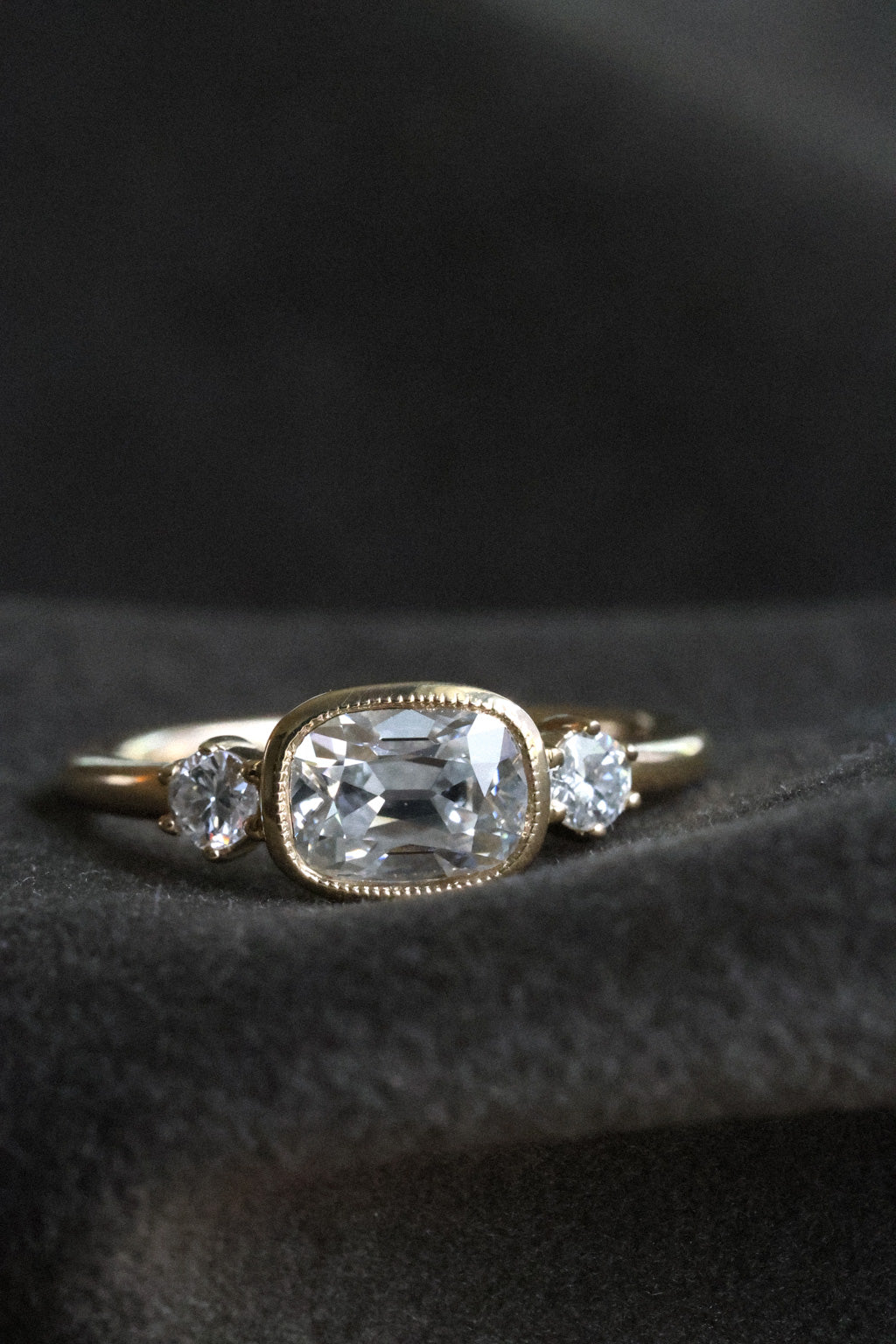 Close up front view photo of an engagement ring resting on dark charcoal wool fabric against a dark background. The ring is a thin yellow gold band. It has an old mine cut diamond set east-west in a milgrain bezel, with a round old cut diamond held in prongs on either side.