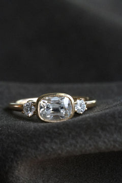 Close up front view photo of an engagement ring resting on dark charcoal wool fabric against a dark background. The ring is a thin yellow gold band. It has an old mine cut diamond set east-west in a milgrain bezel, with a round old cut diamond held in prongs on either side.