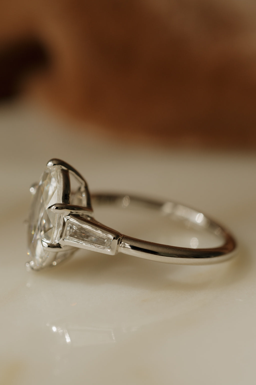 Side profile of a white gold engagement ring with the old mine cut oval diamond facing left, showing a bezel-set tapered baguette diamond and the slim polished band, on a cream surface in warm light.