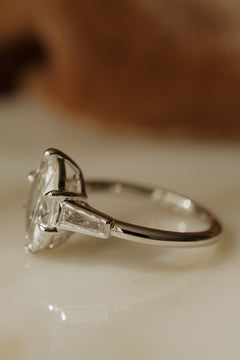Side profile of a white gold engagement ring with the old mine cut oval diamond facing left, showing a bezel-set tapered baguette diamond and the slim polished band, on a cream surface in warm light.