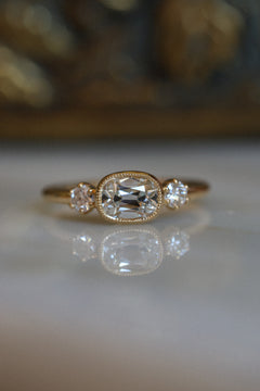 Front view of a yellow gold engagement ring with an old mine cut diamond set east-west in a milgrain-edged bezel, between two round old-cut diamonds in claw prongs, resting on a reflective cream marble surface with an antique-style brass jewelry box blurred behind it.