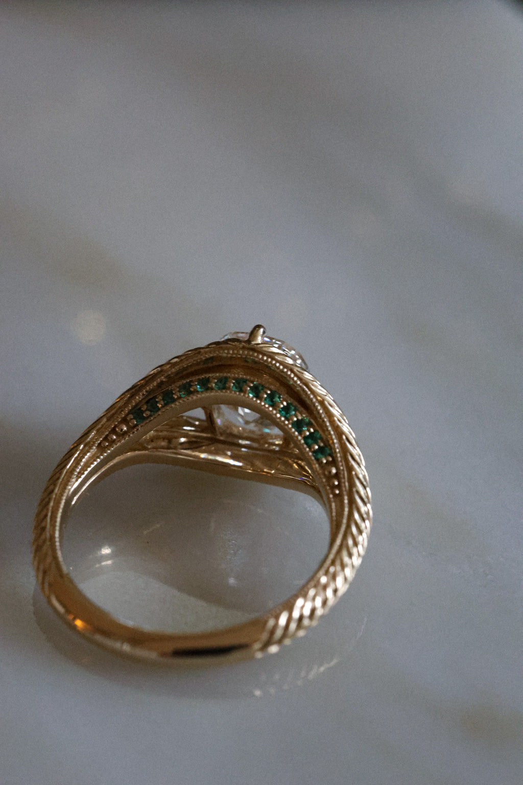 A picture of the underside view of engagement ring, resting on a marble surface. The ring is yellow gold with an old-world hand engraved split shank band. The center stone is an old mine cut diamond with four claw prongs in a compass setting. The split shank has a hidden halo made of emeralds on the lower profile of the side view. 