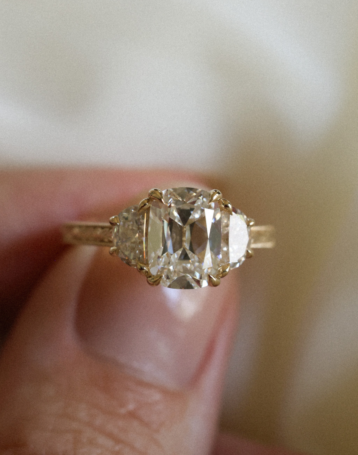 Close view of a fingertip holding a yellow gold three stone engagement ring with an elongated old mine cushion cut diamond in double claw prongs between two half moon diamonds, on an orange blossom engraved band, against soft cream fabric.