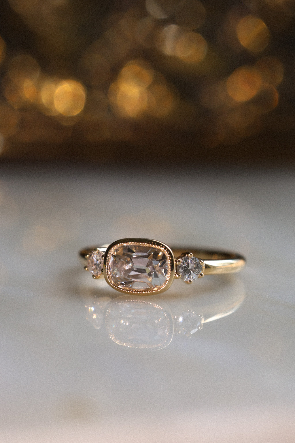 Close up photo of an engagement ring standing on a pale reflective surface, with warm golden lights out of focus behind it and the ring reflected below. The ring is a thin yellow gold band. It has an old mine cut diamond set east-west in a milgrain bezel, with a round old cut diamond held in prongs on either side.