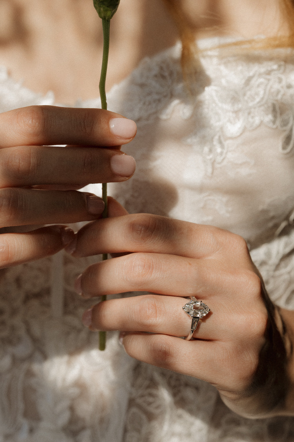 On-hand view of a white gold engagement ring with an old mine cut oval diamond and tapered baguette side stones, the hands holding a single green flower stem in front of a white lace dress.