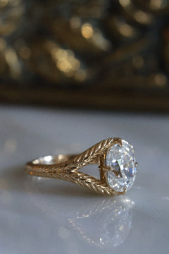 Side view of engagement ring, resting on a marble surface. The ring is yellow gold  with an old-world hand engraved split shank band.  The center stone is an old mine cut diamond with four claw prongs in a compass setting. 