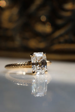Angled front view of a yellow gold engagement ring with an antique emerald cut diamond in four claw prongs and a hand-engraved band, on a reflective cream marble surface with an antique-style brass jewelry box blurred behind.