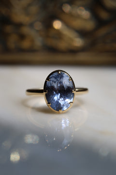 Close up photo of an engagement ring on a white reflective surface, with a gilded jewelry box out of focus in the background. The ring is a thin yellow gold band. It has an oval blue Ceylon sapphire held in a collet setting with eight prongs.