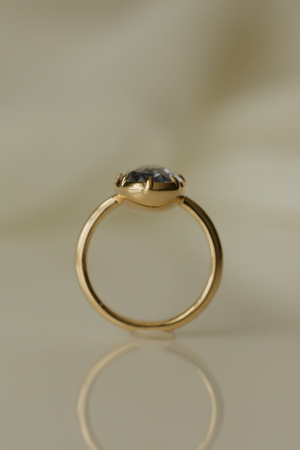 Close up photo of an engagement ring standing upright on a warm beige reflective surface. The ring is a thin yellow gold band. It has an oval blue Ceylon sapphire held in a collet setting with eight prongs.