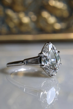 Angled side view of a white gold engagement ring with an old mine cut oval diamond facing right, held by six claw prongs beside a bezel-set tapered baguette diamond, on a white reflective surface before a blurred gilded jewelry box.