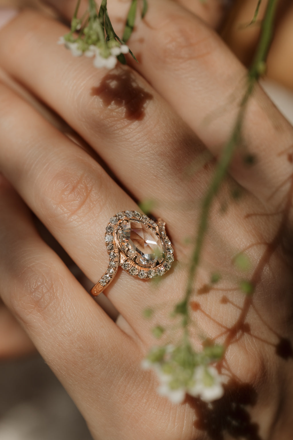 A close up of a hand wearing an engagement ring, there are small white flowers above it. The engagement ring is rose gold and has an oval rose cut diamond with a single cut diamond bypass halo wrapping around it. 