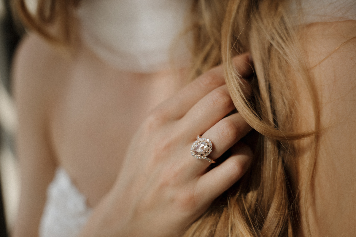 Close view of a hand resting in long strawberry blonde hair, wearing a rose gold engagement ring with an oval rose cut diamond in a curved bypass halo of single cut diamonds, with a bare shoulder and white dress softly blurred behind.