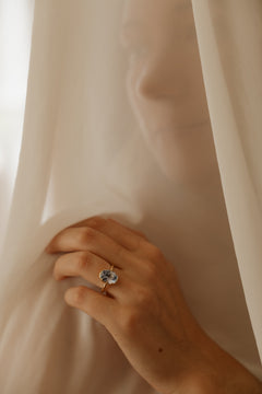 Photo of a hand pressed into a sheer cream curtain, a woman standing behind it, wearing an engagement ring on the ring finger. The ring is a thin yellow gold band with an oval blue Ceylon sapphire held in a collet setting.